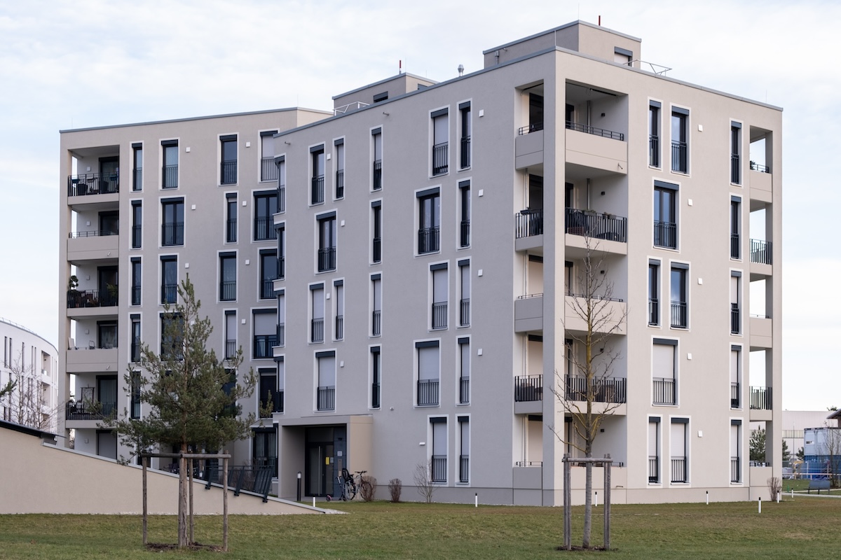 Immeuble moderne de plusieurs étages avec balcons, entouré d'une zone herbeuse et d'arbres épars, sous un ciel partiellement nuageux.