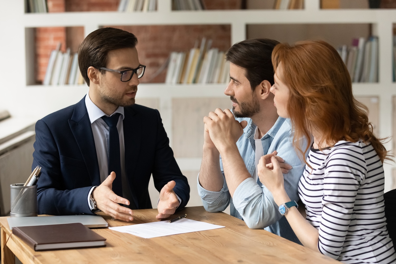 Un homme d'affaires en costume est assis à une table pour discuter de la paperasse avec un couple. Le décor semble être un bureau ou une bibliothèque avec des étagères de livres en arrière-plan.