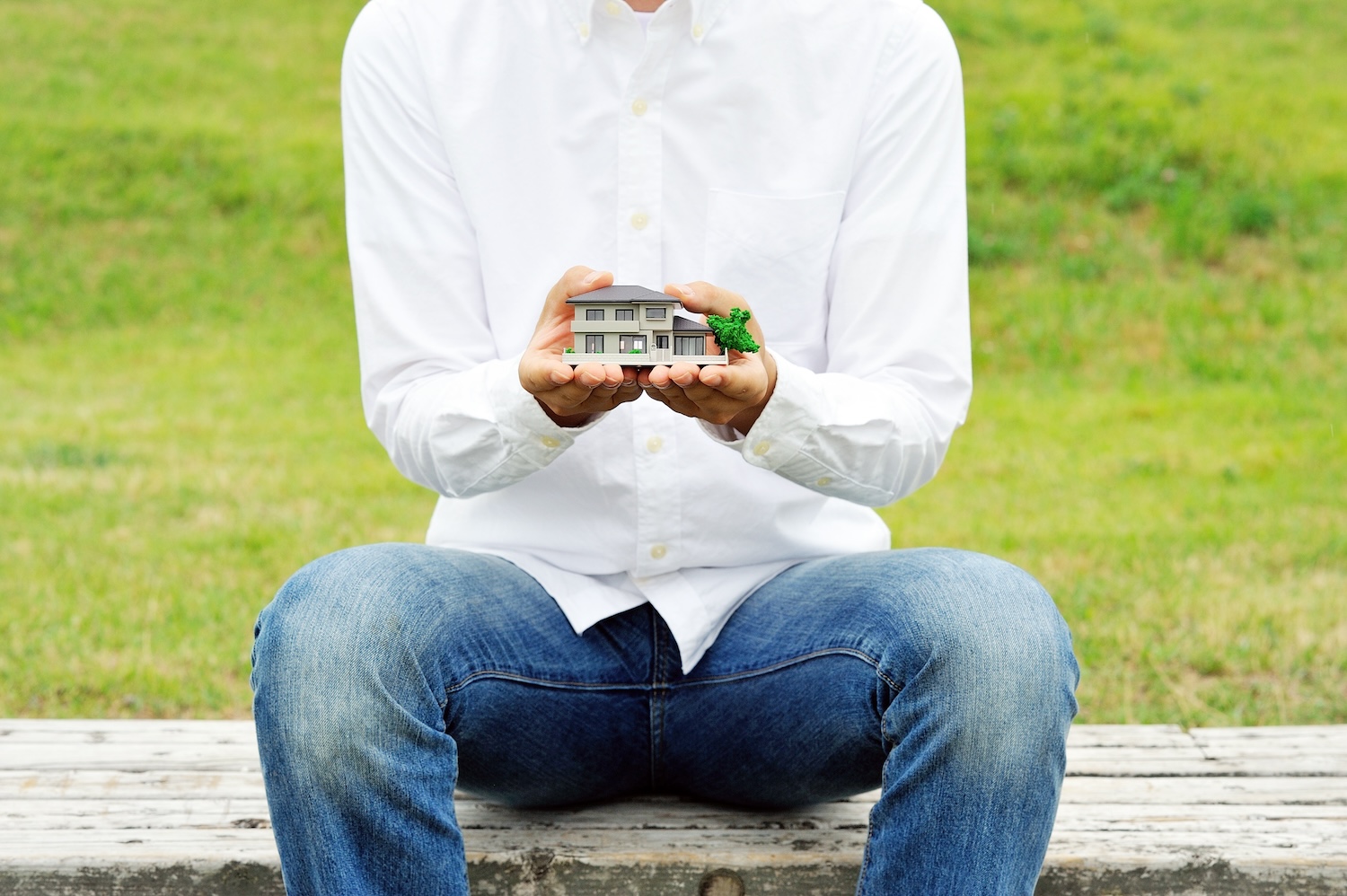 Une personne vêtue d'une chemise blanche et d'un jean bleu tient une petite maison modèle dans ses mains alors qu'elle est assise sur un banc extérieur avec de l'herbe en arrière-plan.