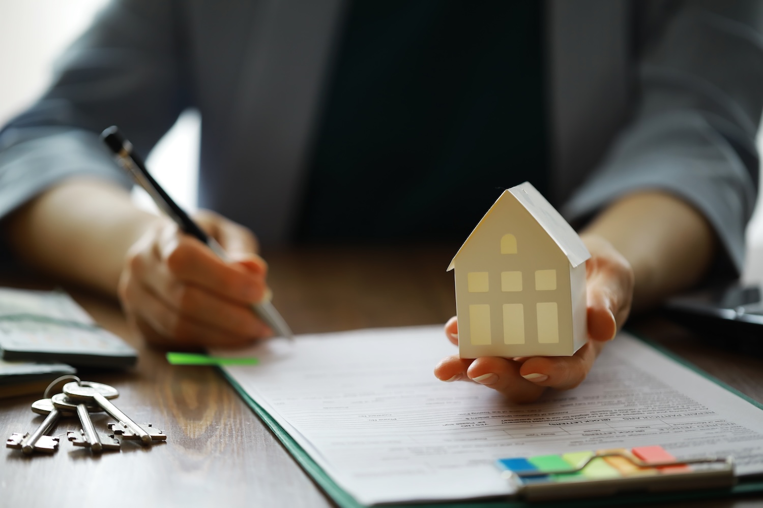 Une personne signe un document en tenant une petite maquette de la Maison Blanche. Des clés, du papier-monnaie et un bloc-notes sont visibles sur la table.