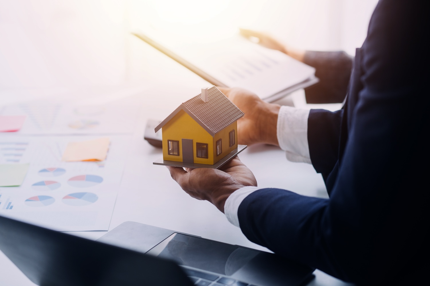 Une personne en costume d'affaires tient un petit modèle de maison jaune tout en examinant des documents et des graphiques à un bureau.