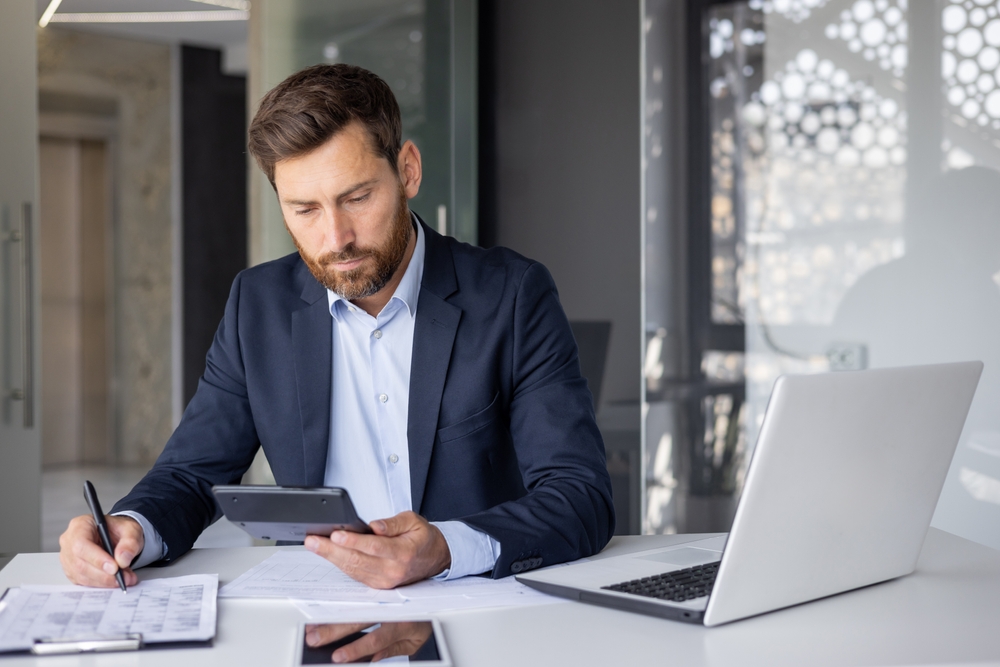 Un homme en costume est assis à un bureau, regardant une tablette tout en écrivant sur des papiers. Un ordinateur portable est ouvert à côté de lui.