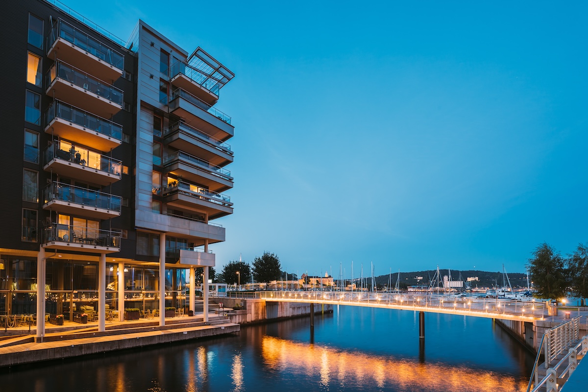 Des immeubles d'appartements modernes et un pont piétonnier éclairé se reflètent dans les eaux calmes d'une marina au crépuscule, avec des bateaux amarrés en arrière-plan.