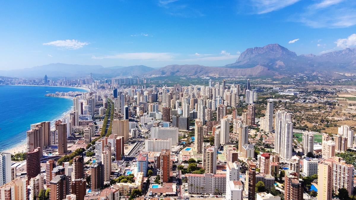 Vue aérienne d'une ville côtière avec de nombreux immeubles de grande hauteur au bord de la mer, entourée de montagnes et d'un ciel bleu clair.