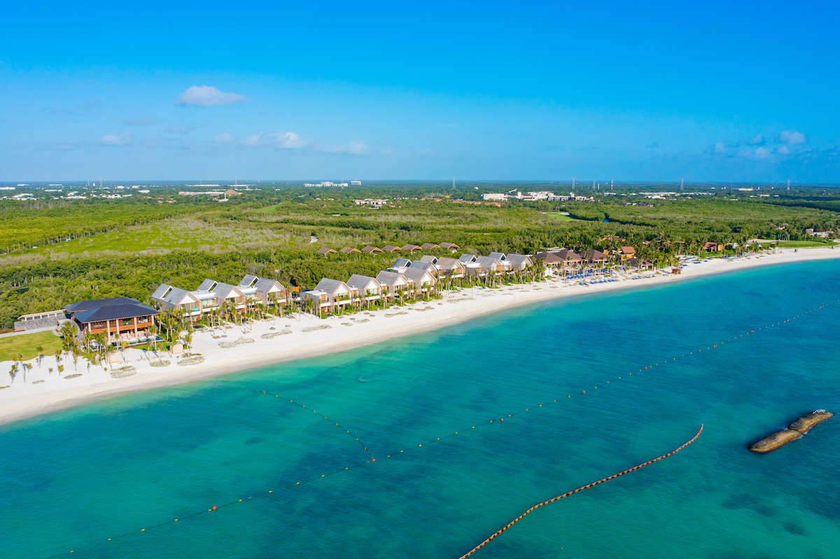 Vue aérienne d'une station balnéaire aux eaux turquoise, plage de sable et végétation verte en arrière-plan sous un ciel bleu clair.
