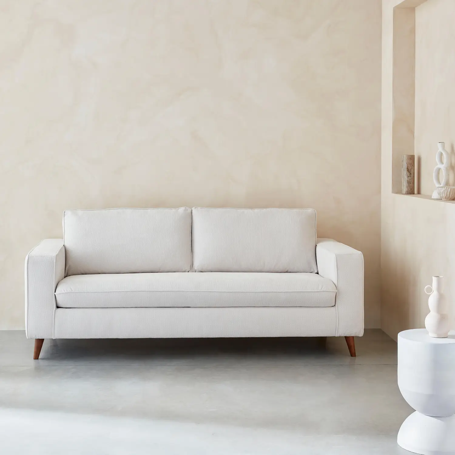 Un salon minimaliste avec un canapé beige, une table d'appoint ronde avec des objets décoratifs et une étagère intégrée avec de petits vases contre un mur aux tons neutres.