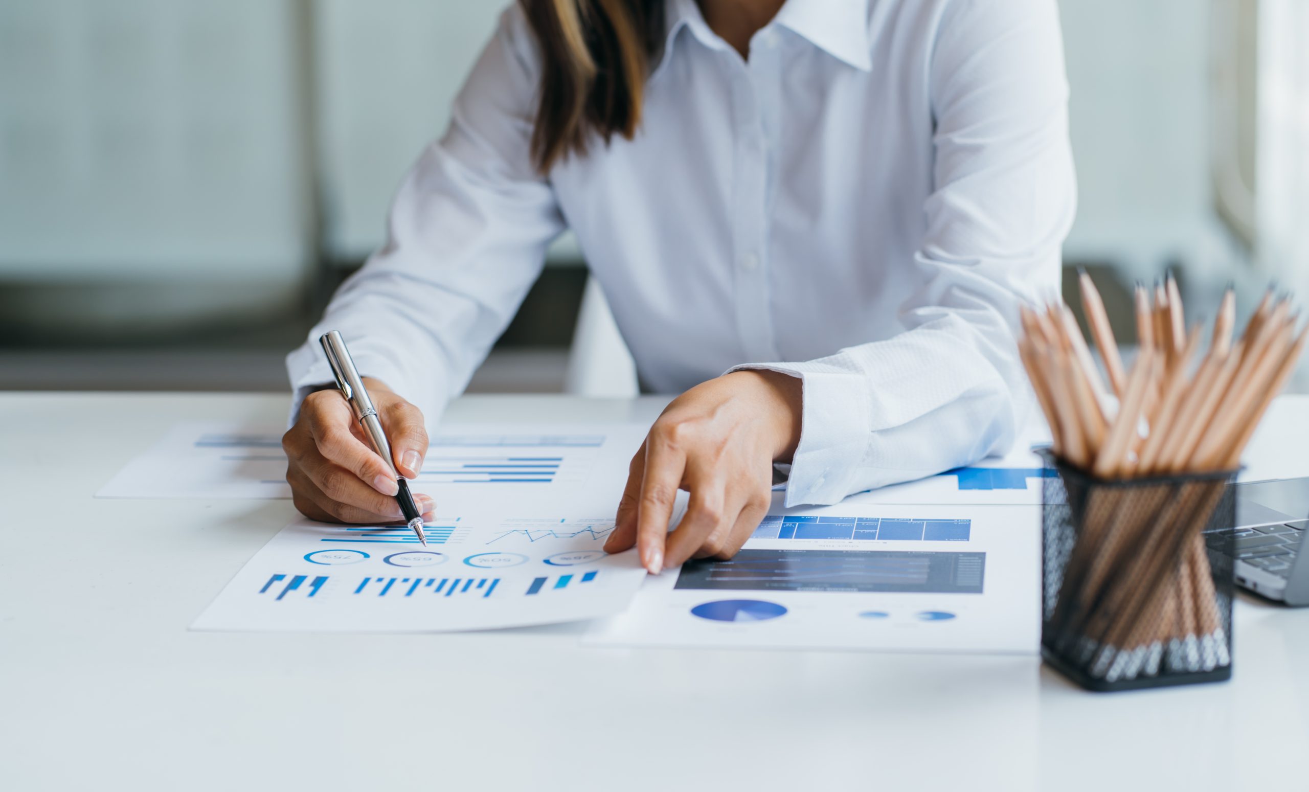 Une personne en chemise blanche analyse des graphiques sur papier, tenant un stylo. Un bureau avec des crayons dans un support est visible à côté d'eux.