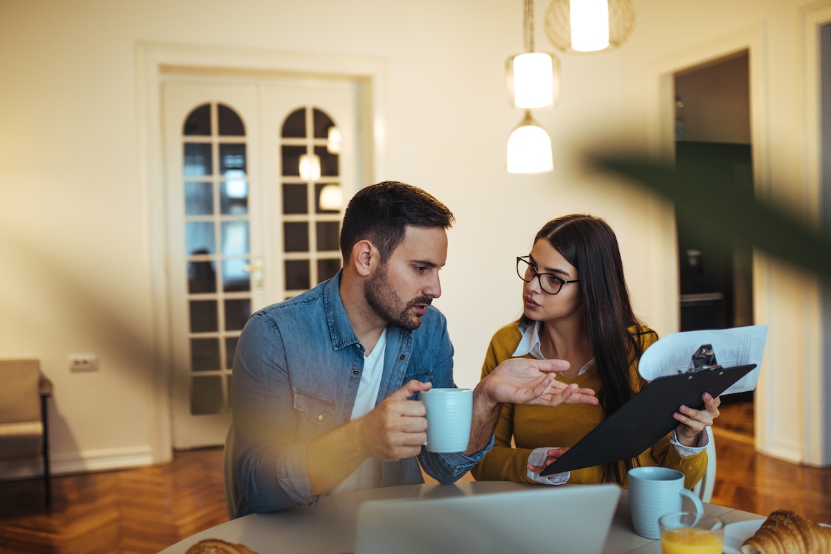 Un homme et une femme sont assis à une table et discutent tout en tenant une tasse et un presse-papiers. Un ordinateur portable et un verre de jus d'orange sont posés sur la table.